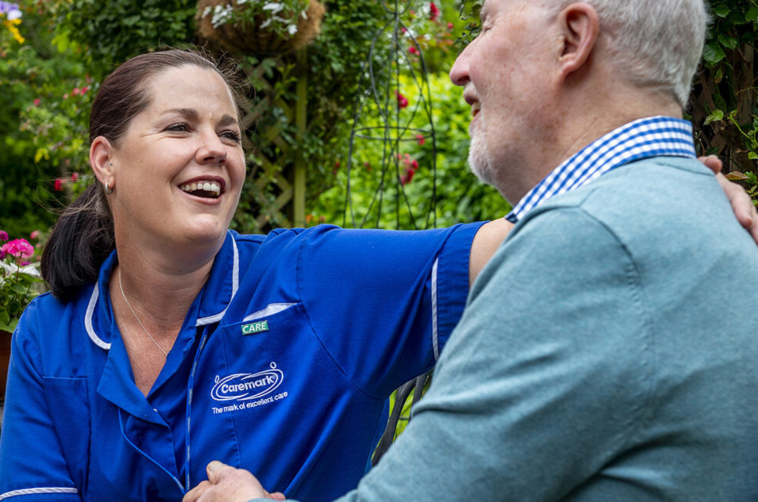 A Caremark carer in a blue uniform smiling with an elderly man in a garden