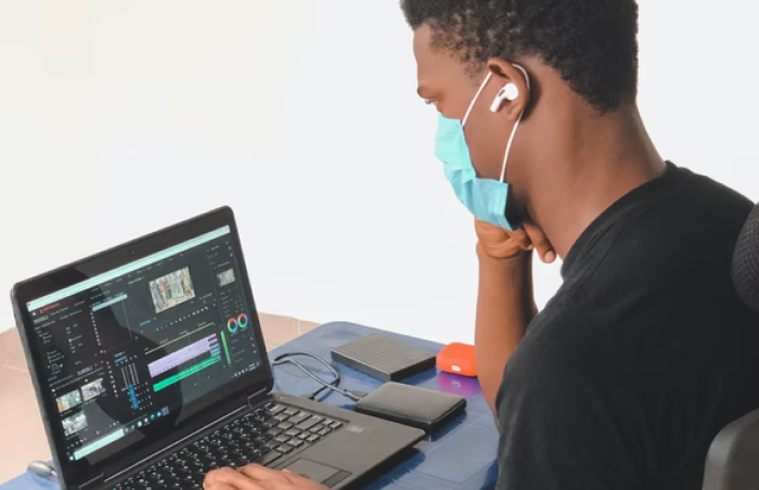 A masked designer editing video and marketing content on a laptop